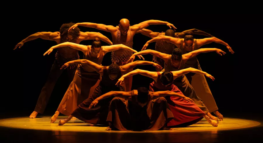 Nine dancers in a cluster looking downward with their arms outstretched in dim yellow lighting.