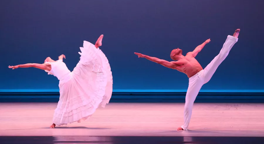 A dancer in a long white dress and a dancer in white pants perform a high kick onstage with their torsos thrown back. 