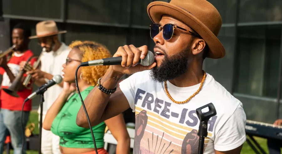 Joe Davis singing in a microphone wearing a white graphic tee and a brown hat, with other performers to his right.