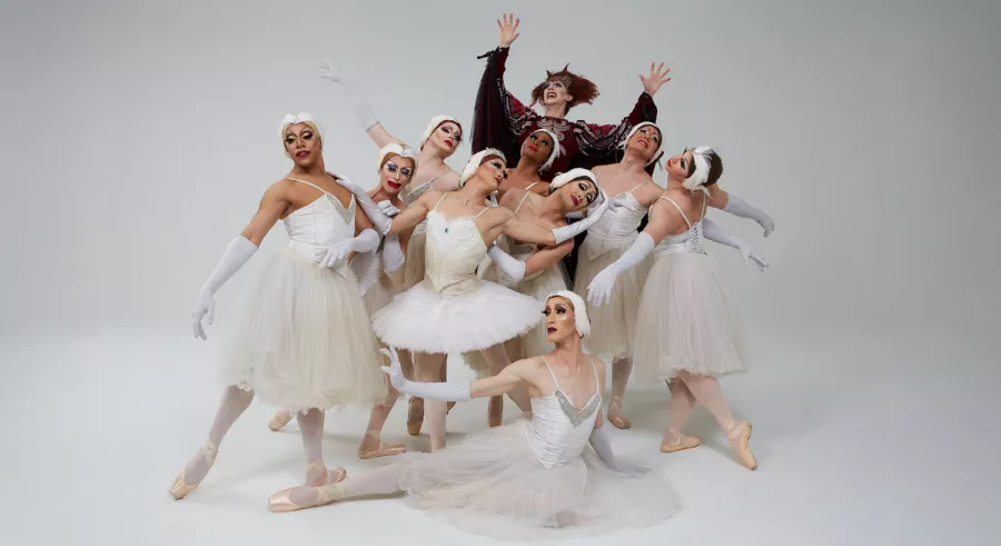 A group of male dancers in flamboyant drag costumes pose in front of a white background.