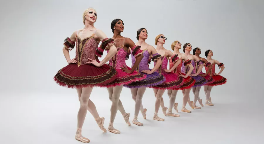A line of dancers in red tutus standing with their right legs bent behind them and their hands on their hips in front of a white background.