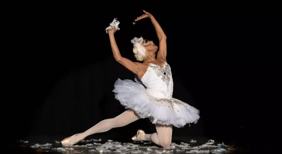 A dancer in a white tutu kneeling with his right leg pointed straight out, looking and holding his arms upwards. 