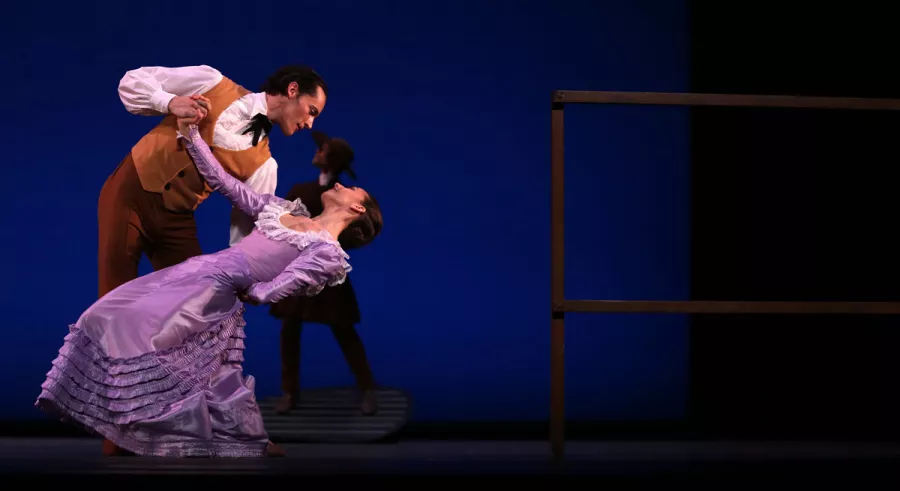 A man in a brown vest dips a woman in a long purple dress onstage with someone standing behind them in shadow.