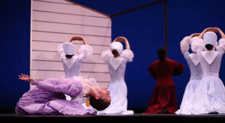 A dancer in a purple dress kneeling and bending backward while four dancers in light blue dresses and a dancer in a red dress kneel behind her, facing away.