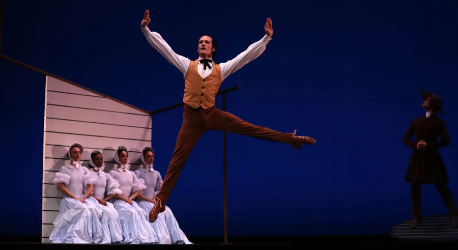 A dancer in a brown vest and brown pants leaping while four dancers sit behind him and another dancer stands off to the side. 