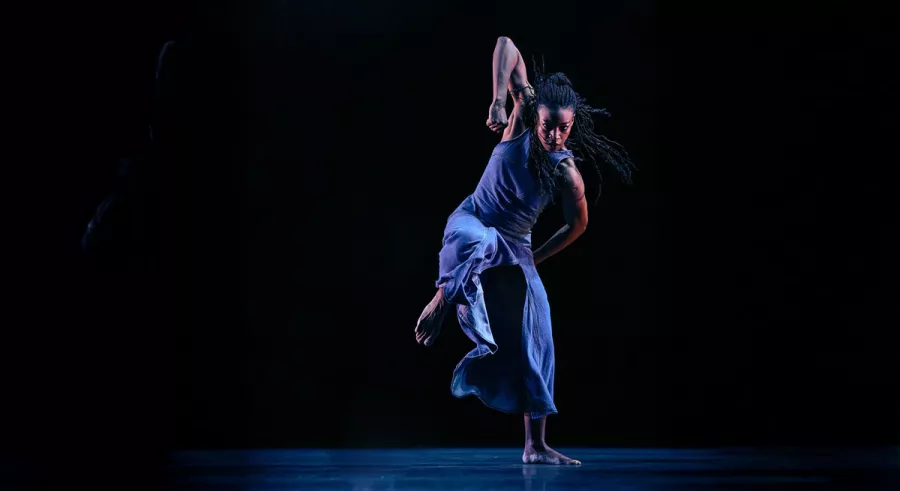 A dancer poses with both arms up and one leg bent in front of a black background. 