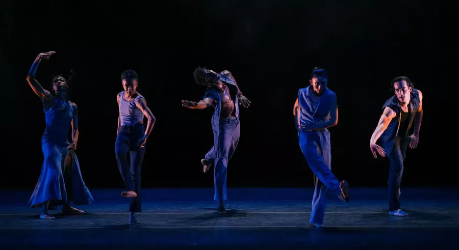 Five dancers in a line performing onstage in front of a dark background. 