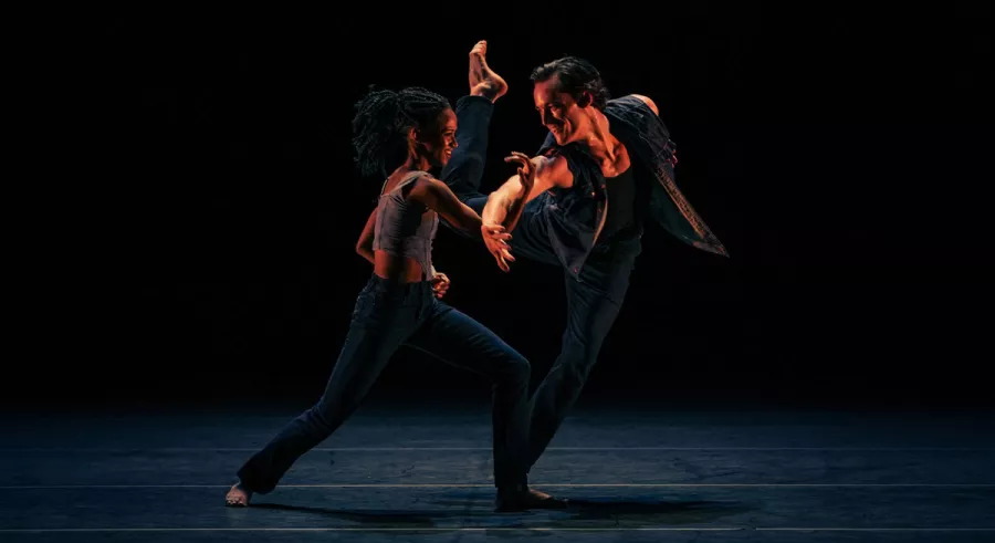 Two dancers right next to each other in differing poses in front of a dark, foggy background.