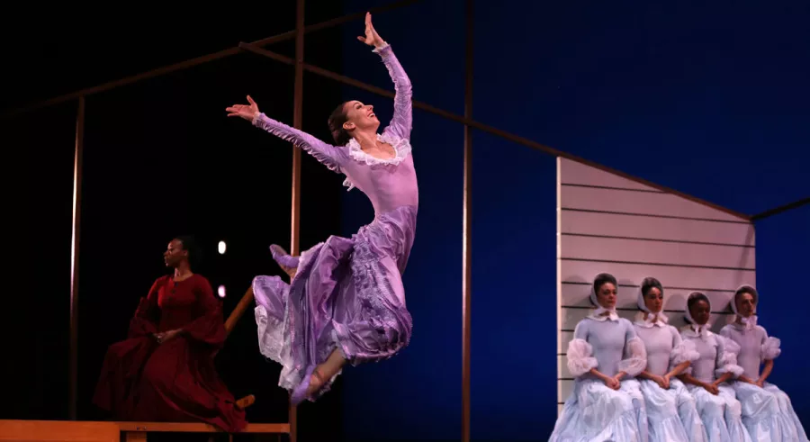 A dancer in a purple dress leaping onstage with four dancers in light blue dresses and another dancer in a red dress sitting behind her.