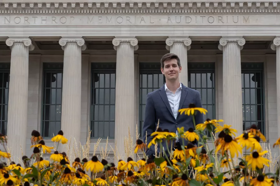 Greg Zelek, a man in a navy suit, stands in front of the Northrop Auditorium by yellow flowers.