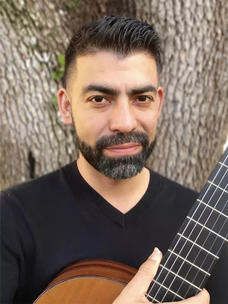 Alvaro Bermudez outdoors with his guitar by a tree.