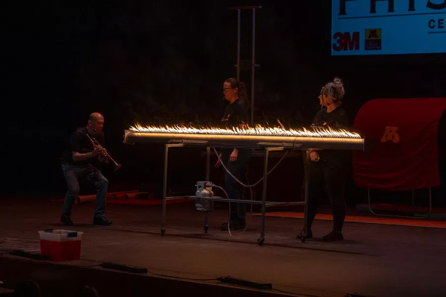 Two people stand behind a rod of flames and a man holding a trumpet crouches at one end.