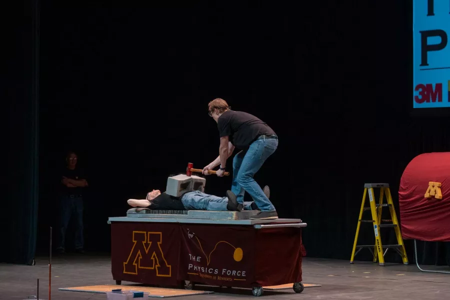 A man lies on bed of nails with a cinder block on his torso, another man is hitting the block with a sledgehammer.