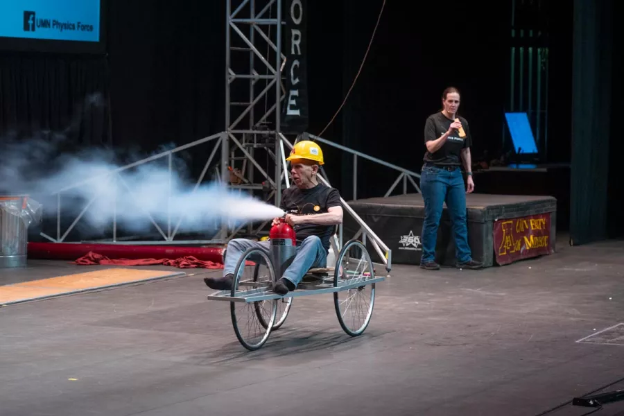Man sits in a tri-wheeled chair holding a discharging fire extinguisher in his lap.
