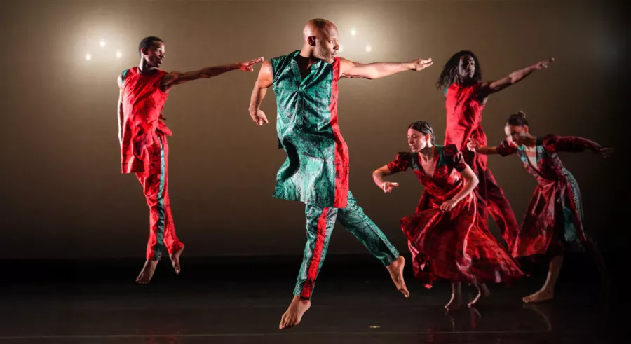 Five dancers in red and green outfits performing on stage, captured mid-motion.