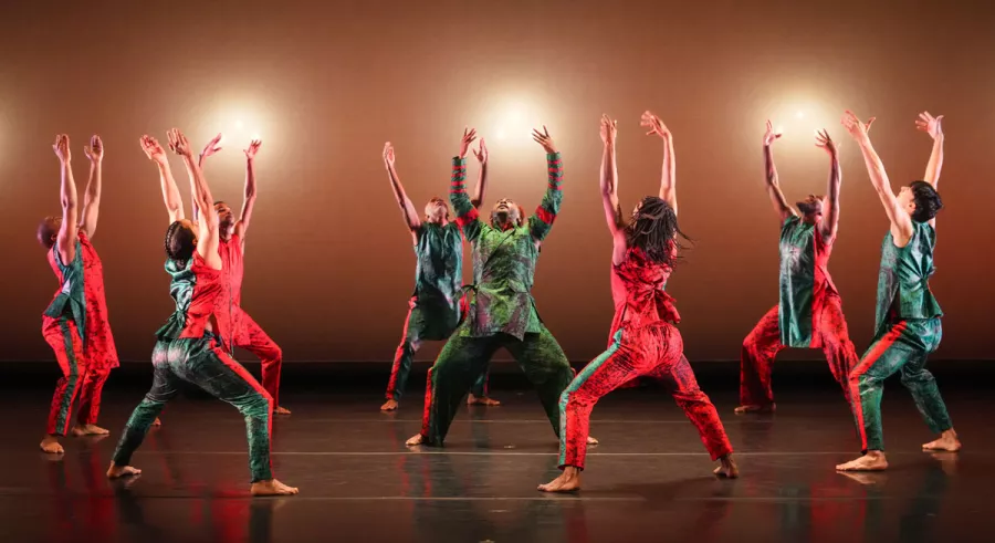 A group of nine dancers performing on stage in red and green costumes, reaching upwards against a warm background.