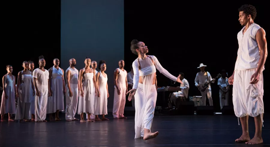 Ronald K. Brown company members dressed in white. Left: Company standing straight in a clump. Right: One dancer with arms infront. Other dancer standing from a distance. 