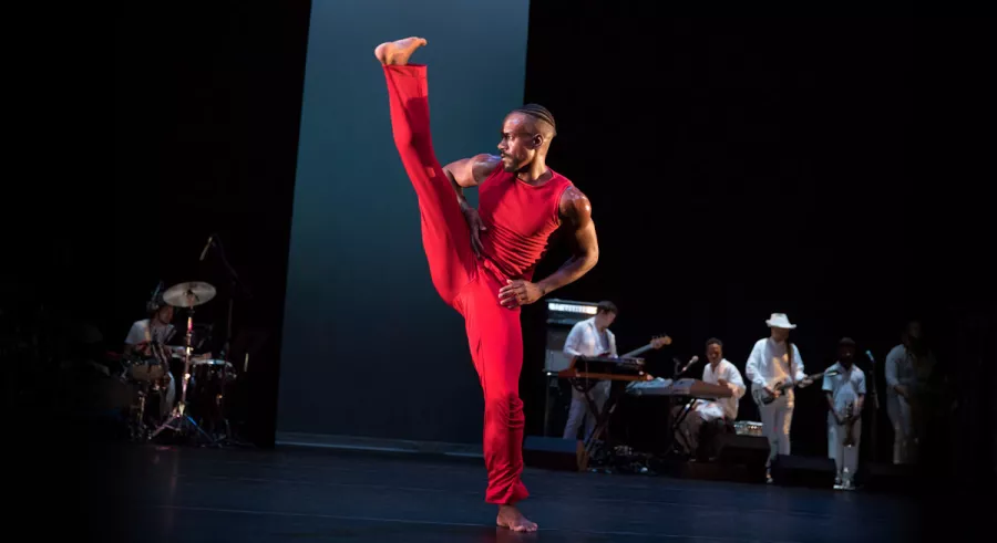 Dancer in red pants and sleevles top kicks leg upward with hands on hips.