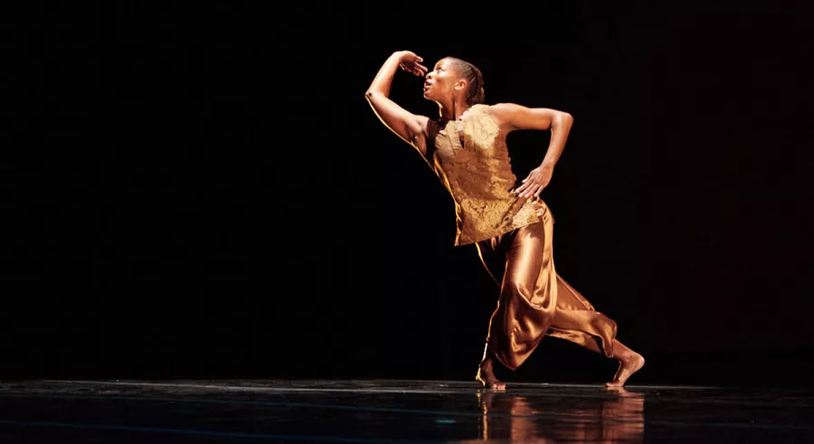 Dancer wearing a gold is captured in a dynamic pose, with one arm bent gracefully over their head and the other positioned on their hip.