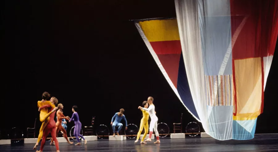 Dancers in an array of colorful outfits perform onstage next to a large, patchwork curtain.