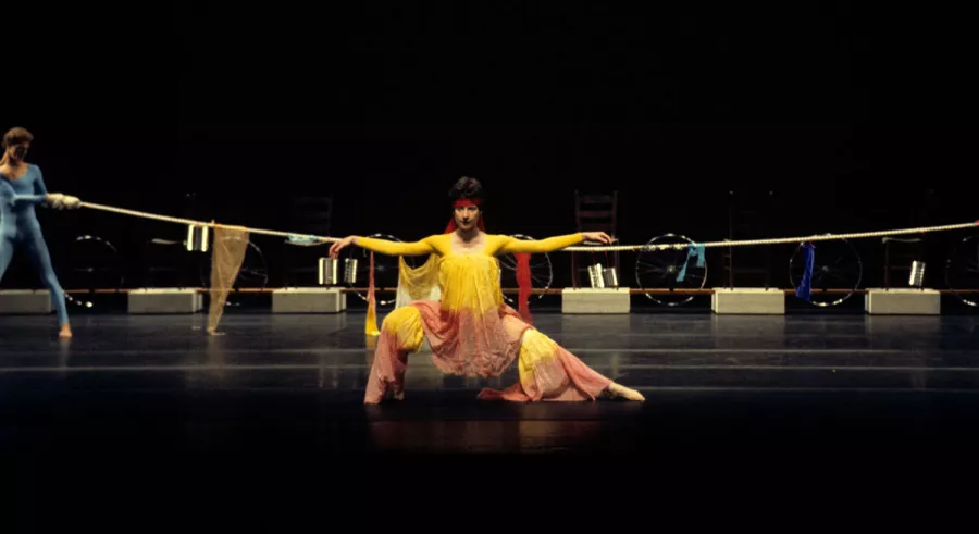 A dancer in a yellow outfit crouching low to the ground with both arms outstretched onstage. 