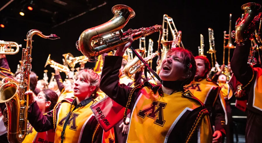 A close-up on a saxophone player raising his instrument while cheering amidst the rest of the band doing the same. 