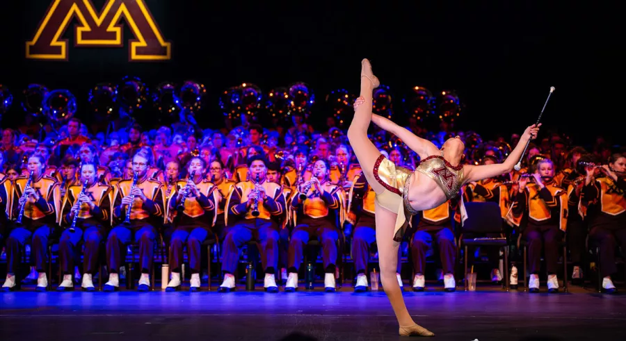 A baton twirler doing a high kick while catching a baton in front of the entire UMN Marching Band. 