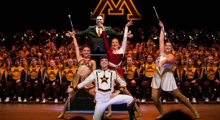 Drum major Ari Martin, the UMN Feature Twirlers, and Assistant Director John Leonard pose in front of the UMN Marching Band after a performance. 