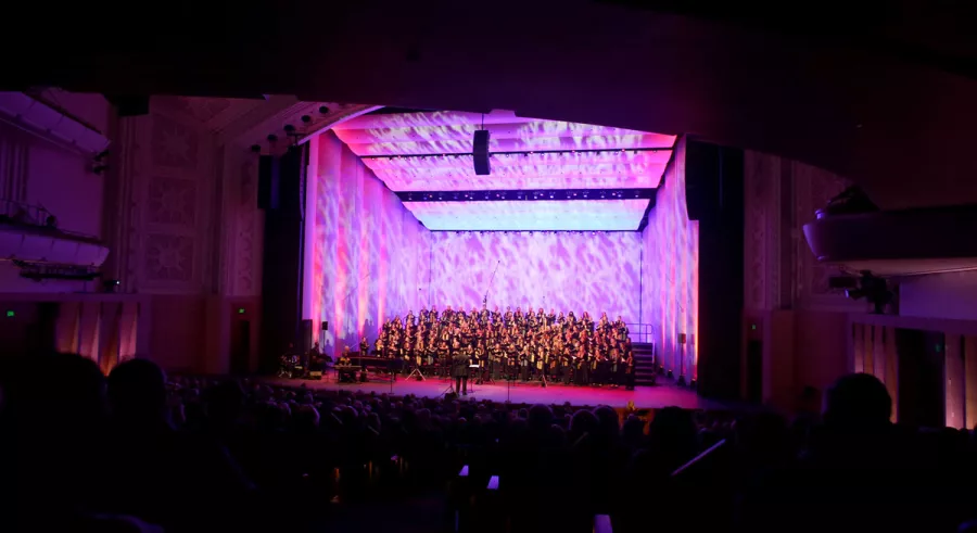 A picture from the back of the audience of the singers of the WITNESS choir performing onstage with a conductor at Northrop with a purple and pink spotted background.