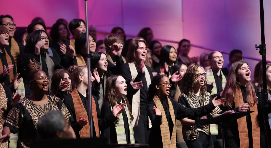 Members of a choir singing onstage wearing stoles in front of a purple background.