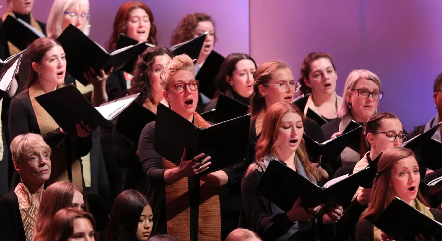 A choir sings with music in their hands in front of a purple background.