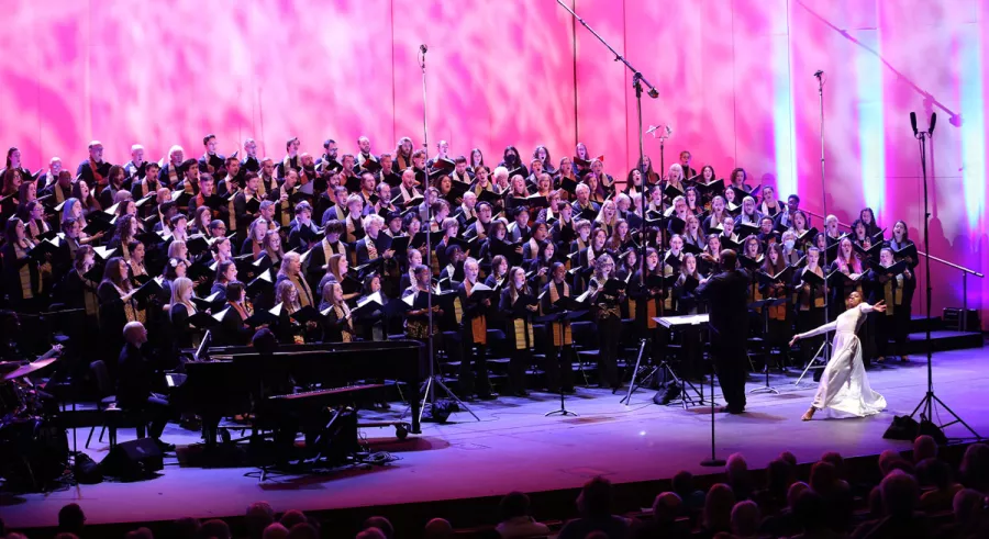 A choir performs in front of a pink background with a woman in a white dress dancing in front of them.