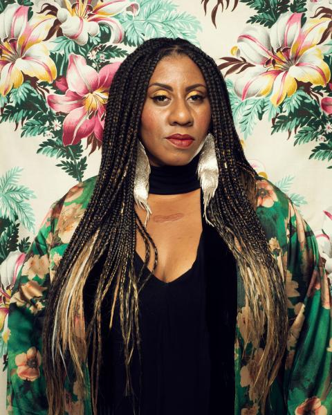 Annie Mack, a black woman with long braided hair, wears a black top, with a green silk flowered jacket, and large silver feather earrings. Large floral print background.