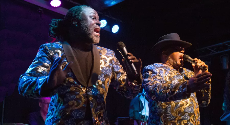 2 men sing into microphones on a lit stage. They are both black men wearing decorative jackets, the man on the right is wearing a black brimmed hat, the man on the left has his hair tied back.