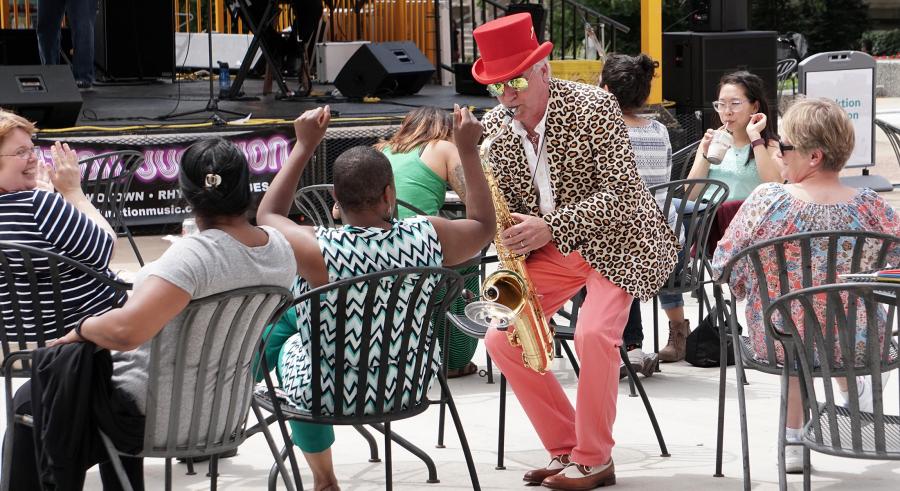 Saxophonist plays in the crowd. He is a white man wearing salmon colored pants and top hat, leopard print jacket.
