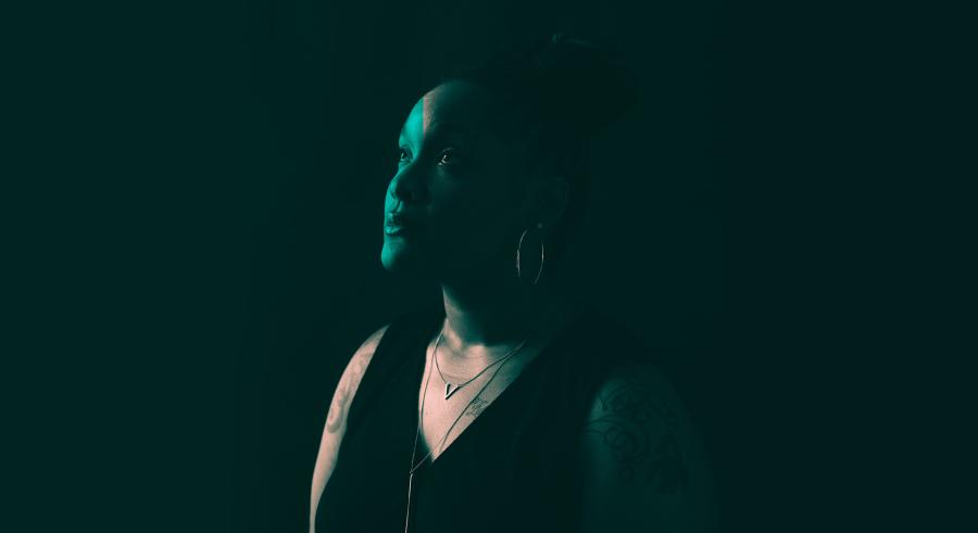 PaviElle, a black woman wears a black tank dress, her hair is pulled back and tattooed shoulders, looks up into blue-green light that shines on her face