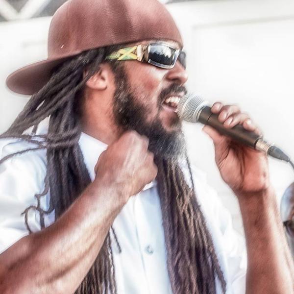 Jabba, a black man with a beard and long braided hair, sings into a microphone. He is wearing a brown ball cap backwards, gold sunglasses and a white button-down shirt.