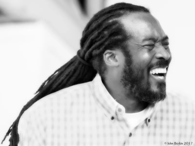Black and white image of Jabba with a laughing smile, wearing a checked shirt, braided hair tied in the back.