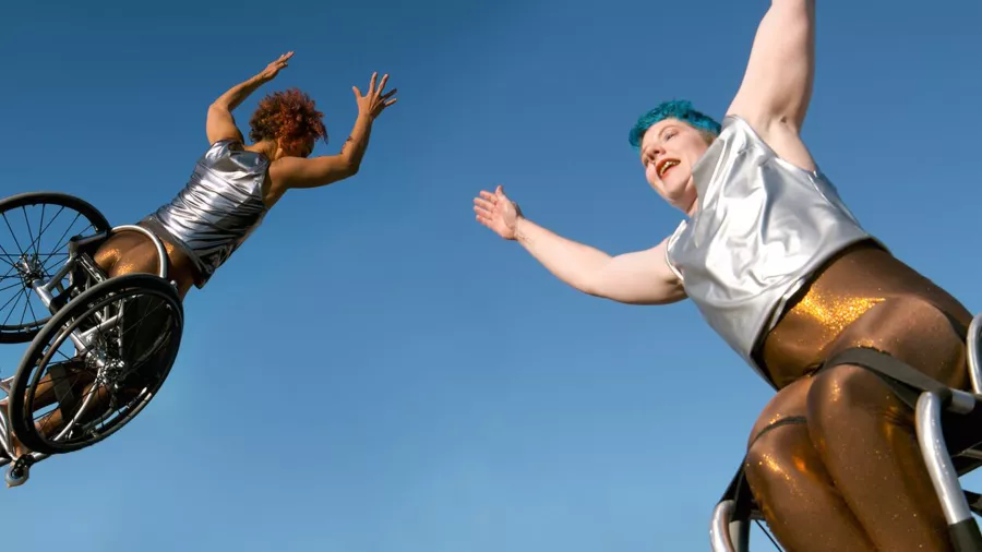 Two dancers in wheelchairs flying through the air against a blue sky