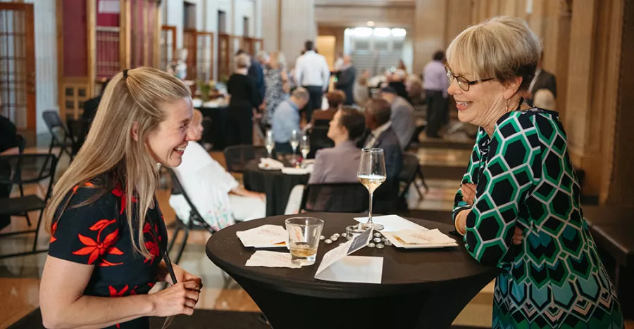 Two women with blonde hair stand at a black circular table. The woman on the right is wearing a black dress with red flowers and the woman on the right is wearing a black and blue dress. Both women are smiling.