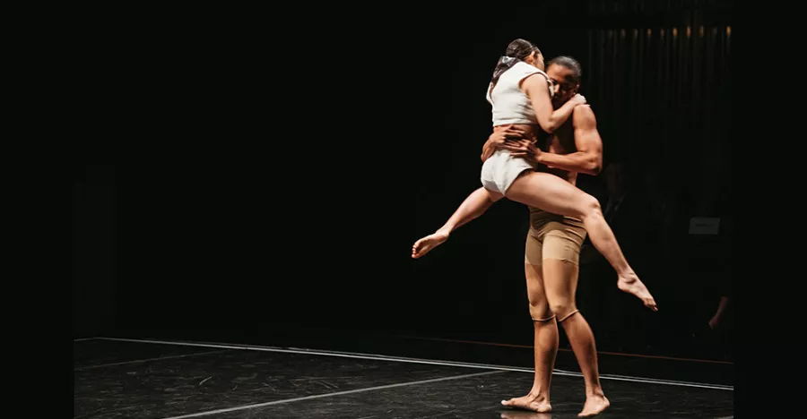 Two dancers are in front of a black background. The dancer on the right is holding the dancer on the left whose legs are extended out to the sides.