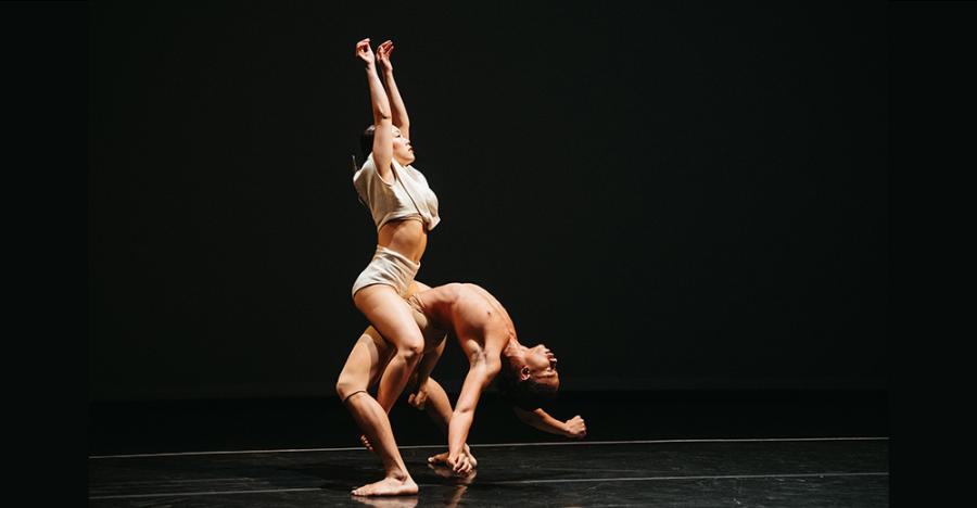 Two dancers are in front of a black background. One dancer is standing with their back arched. The other dancer is on top of them with their arms raised.