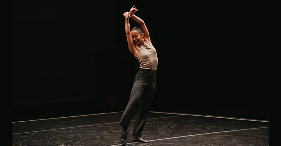 A dancer stands on a black floor in front of a black background. They are standing with their right leg bent so their toe is touching the ground. They are wearing black pants and a beige shirt.