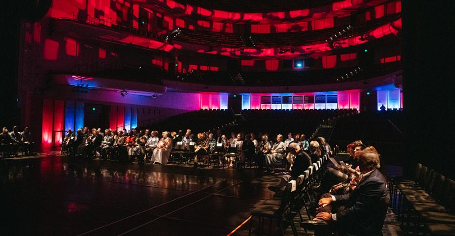 Audience members sit in black chairs surrounding the stage. In the background, the theater has red and blue lights projected on the seats and walls.