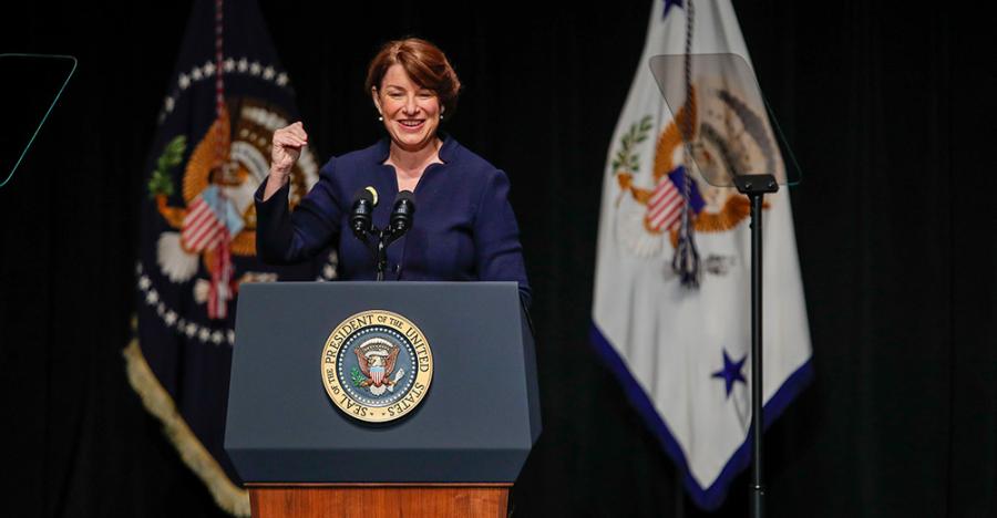 Senator Amy Klobuchar with brown hair wearing a blue blazer stands behind a podium on stage. The podium has the presidential logo on the front side, and two flags hang behind her. 
