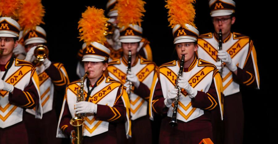 A group of marching band members wear matching maroon, gold, and white uniforms, all playing flutes and saxophones. In the lower right of the photo is a yellow urn, and an american flag, folded within a triangular wooden and glass case.
