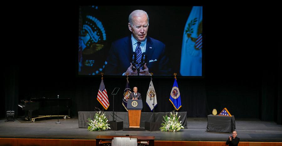 President Biden wearing a blue suit stands behind a podium on stage. The podium has the presidential logo on the front side, and four flags hang behind him. 