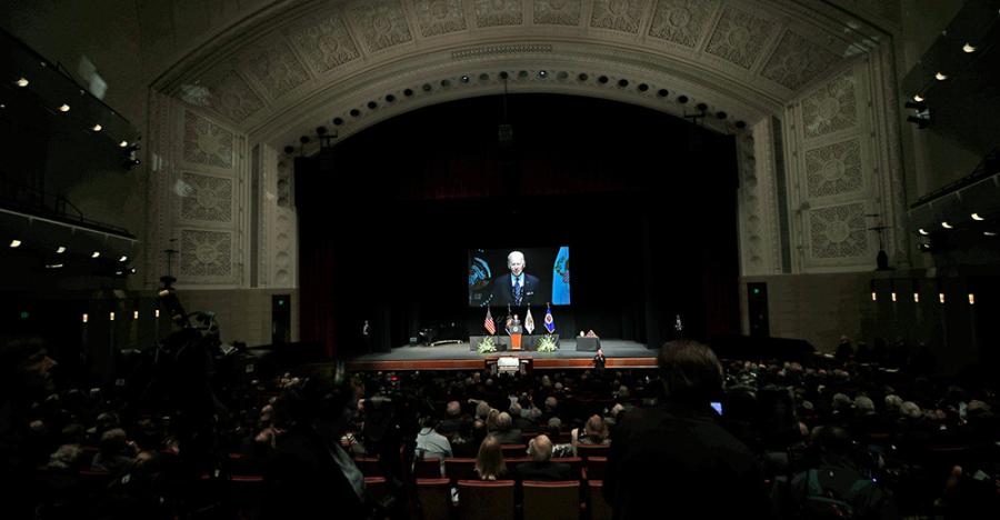 Northrop theater filled with patrons paying their respect to Walter Mondale. The screen shows President Biden delivering a speech with four flags behind him. 
