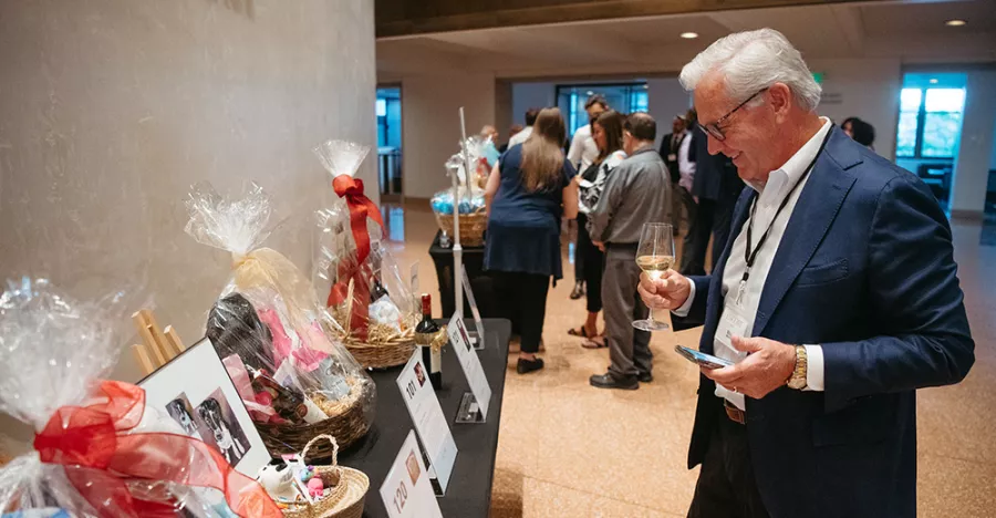 A person wearing a blue suit jacket holding a phone in their left hand and glass of white wine in their right reads a white paper in front of gift baskets.