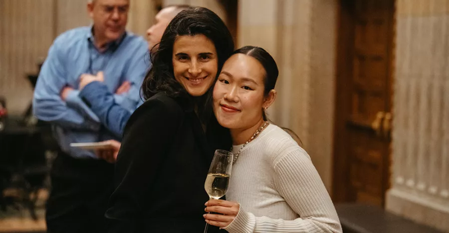 Two women pose for a picture in front of cream colored walls and brown doors. The woman on the left has her hair down and is wearing black. The woman on the right has her hair pulled back and is wearing white.